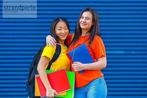 Porträt von zwei niedlichen multiethnischen Studentinnen  die sich vor einem blauen Hintergrund umarmen