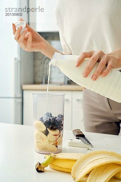 Unbekannte Frau gießt Milch aus der Flasche in die Schüssel des Mixers  um einen Smoothie mit schwarzen Johannisbeeren herzustellen