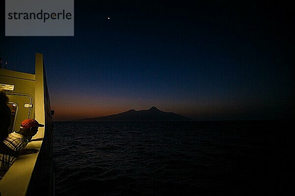 Mann steht an einer Reling mit einem roten Hut und schaut auf die Silhouette einer Insel kurz nach Sonnenuntergang