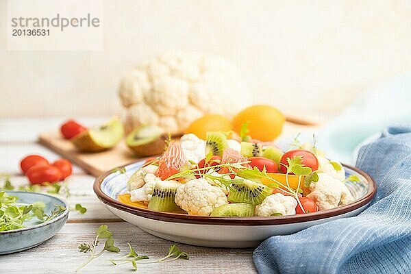 Vegetarischer Salat aus Blumenkohl  Kiwi  Tomaten  Mikrogrünsprossen auf weißem Holzhintergrund und blauem Leinentuch. Seitenansicht  Nahaufnahme  selektiver Fokus