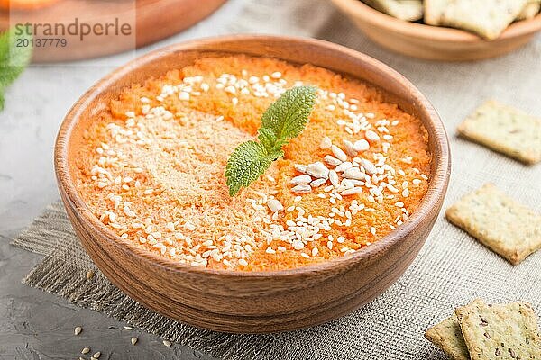 Karottencremesuppe mit Sesam und Knabbereien in Holzschale auf grauem Betonhintergrund mit Leinentextil. Seitenansicht  Nahaufnahme  selektiver Fokus