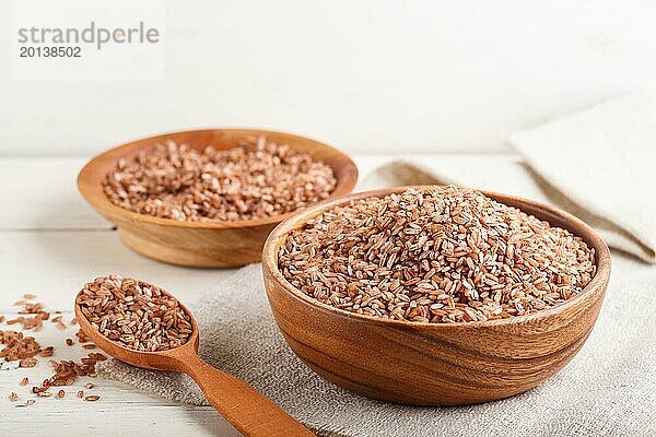 Zwei Holzschalen mit ungeschliffenem braunem Reis und Holzlöffel auf weißem Holzuntergrund und Leinenstoff. Seitenansicht  Nahaufnahme  selektiver Fokus