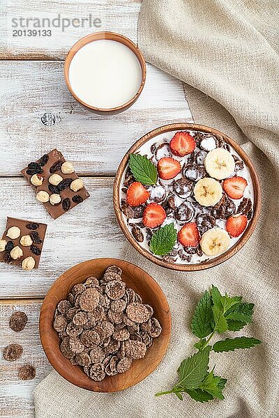 Schokoladen Cornflakes mit Milch und Erdbeeren in einer Holzschale auf weißem Holzhintergrund und Leinenstoff. Draufsicht  Flachlage  Nahaufnahme