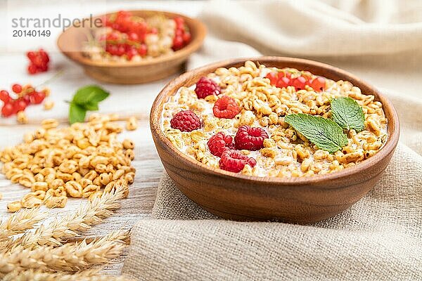 Weizenflockenbrei mit Milch  Himbeeren und Johannisbeeren in einer Holzschale auf weißem Holzhintergrund und Leinentextil. Seitenansicht  Nahaufnahme  selektiver Fokus