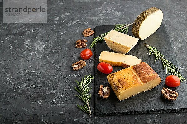 Geräucherter Käse und verschiedene Käsesorten mit Rosmarin und Tomaten auf einer schwarzen Schiefertafel auf einem schwarzen Betonhintergrund. Seitenansicht  Kopierraum  selektiver Fokus