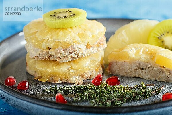 Stücke von gebackenem Schweinefleisch mit Ananas  Käse und Kiwi auf grauem Teller und blauem Hintergrund  Seitenansicht  Nahaufnahme  selektiver Fokus