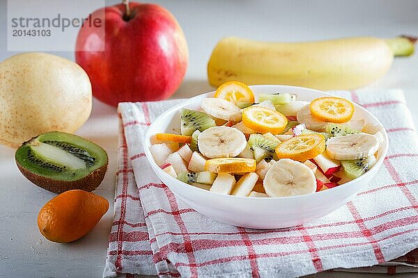 Vegetarischer Salat aus Bananen  Äpfeln  Birnen  Kumquats und Kiwi auf Leinentischdecke  Nahaufnahme  selektiver Fokus. weißer Holzhintergrund