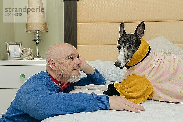 Ein erwachsener Mann betrachtet liebevoll sein Haustier  einen Windhund  der ein orangefarbenes Fell trägt  um sich warm zu halten  während eines spielerischen Moments auf dem Bett