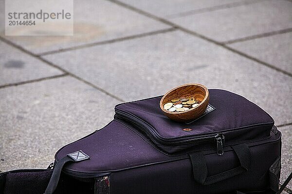 Auf dem Bürgersteig liegender schwarzer Koffer eines Straßenmusikers und eine mit Münzen gefüllte Schale