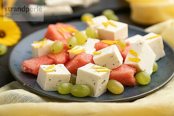 Vegetarischer Salat mit Wassermelone  Fetakäse und Trauben auf blauem Keramikteller auf schwarzem Betonhintergrund und gelbem Leinentuch. Seitenansicht  Nahaufnahme  selektiver Fokus