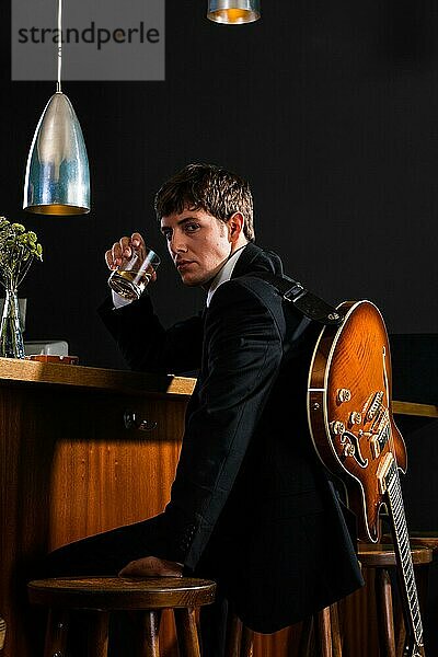 Junger Mann im Anzug sitzt mit seiner Gitarre an einer Bar und trinkt Alkohol