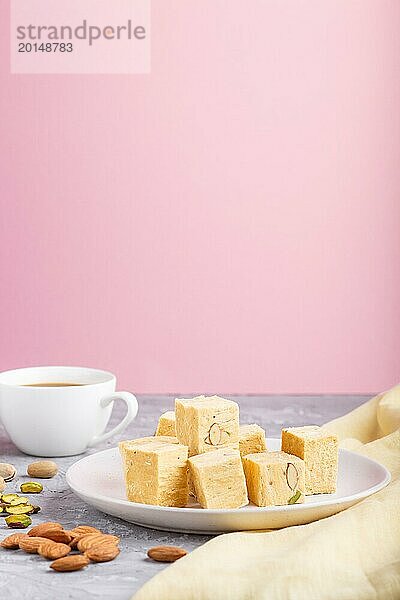 Traditionelle indische Süßigkeit soan papdi in weißem Teller mit Mandel  Pistache und eine Tasse Kaffee auf einem grauen und rosa Hintergrund mit gelbem Stoff. Seitenansicht  Nahaufnahme  selektiver Fokus