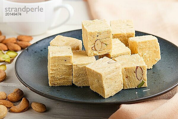 Traditionelle indische Süßigkeit Soan Papdi in einem blauen Keramikteller mit Mandel  Pistache  Cashew und einer Tasse Kaffee auf einem weißen Holzhintergrund mit orangefarbenem Stoff. Seitenansicht  Nahaufnahme  selektiver Fokus