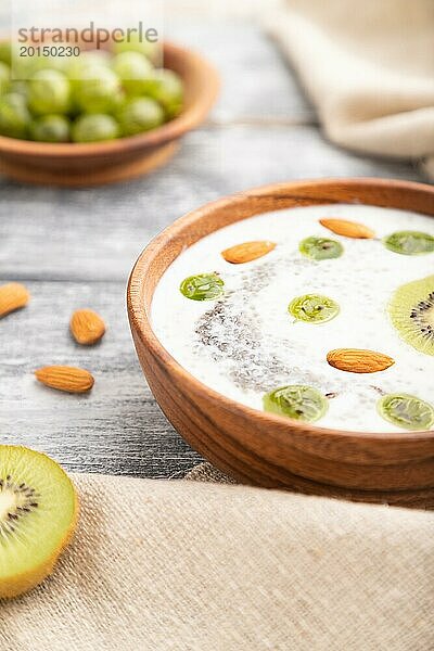 Joghurt mit Kiwi  Stachelbeere  Chia und Mandeln in einer Holzschale auf grauem Holzhintergrund und Leinenstoff. Seitenansicht  Nahaufnahme  selektiver Fokus