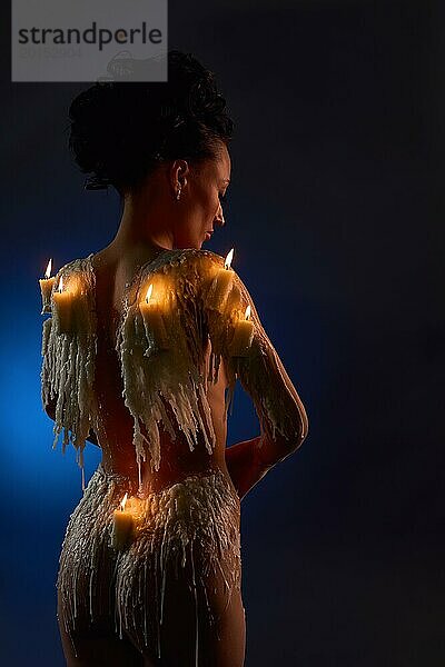 Rückenansicht eines jungen nackten weiblichen Modells mit brennenden Kerzen und Wachs  das den nackten Körper in der Dunkelheit bedeckt