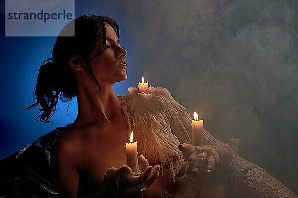 Charmante emotionslose Frau mit brennenden Kerzen in den Händen und auf der Schulter  die vor einem blauen Hintergrund mit Rauch wegschaut