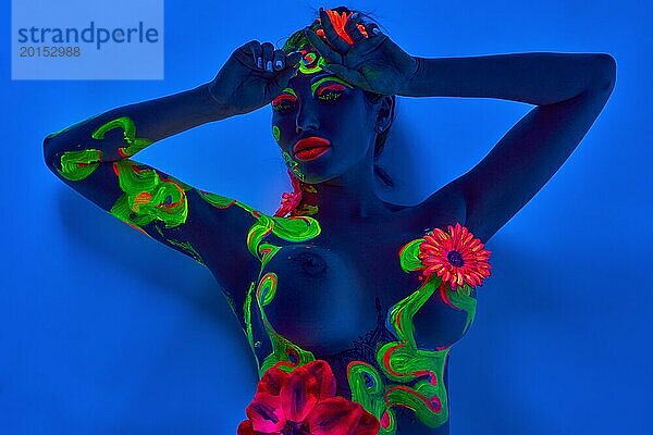 Draufsicht auf nacktes junges weibliches Modell mit Neonmustern und Blumen  das den Kopf berührt und auf blauem Boden liegt
