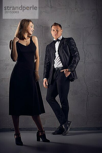 Ganzkörperaufnahme einer zufriedenen Frau in schwarzem Kleid und hochhackigen Schuhen  die gegen ihren Freund in einem eleganten Anzug an einer grauen Wand steht