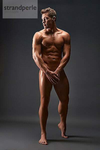 Ganzkörper verführerischen nackten männlichen Modells mit perfekten Torso stehen gegen schwarzen Hintergrund mit den Händen auf Leiste und Blick weg im Studio