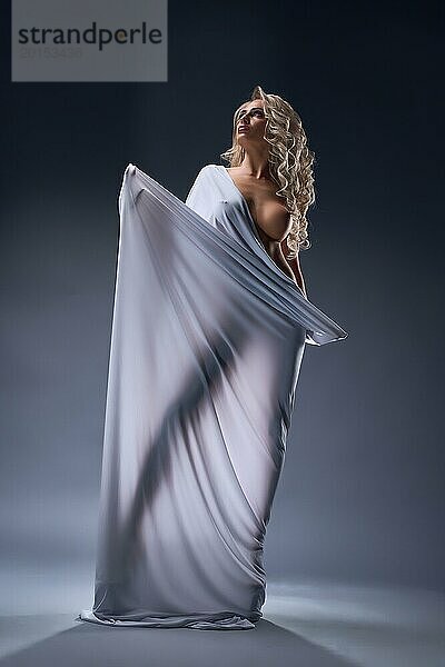 Full Body of charmante nackte weibliche Modell mit langen lockigen blonden Haaren in weißem Tuch gewickelt Blick weg gegen dunklen Hintergrund