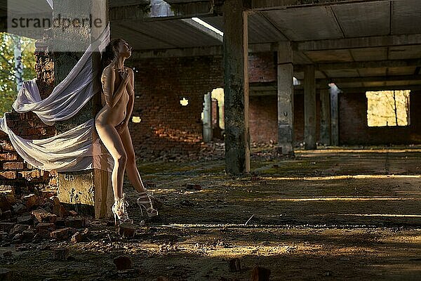Seitenansicht Ganzkörperansicht einer verführerischen nackten Frau  die sinnlich in der Nähe von Betonmasten in einem verlassenen Gebäude mit weißem Stoff steht