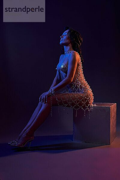 Junges verführerisches weibliches Modell in transparentem Kleid und High Heels sitzt auf einem Würfel in einem dunklen Studio  das von blauen und lila Neonlichtern beleuchtet wird