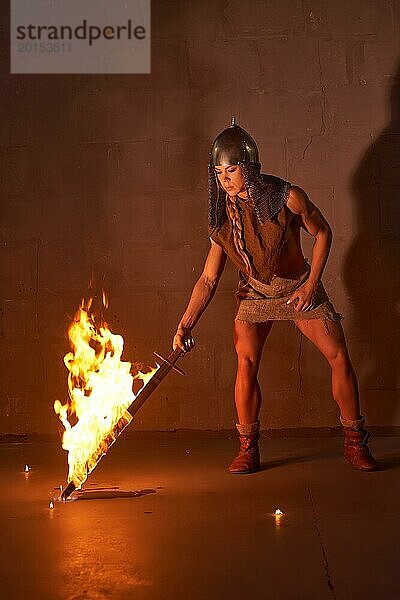 Ganzkörper starke Frau in Wikingerkostüm und Helm trägt brennendes Schwert vor Kampf gegen braune Wand bei Nacht