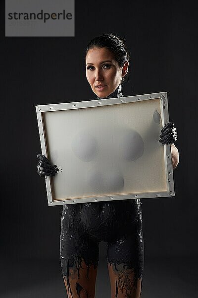 Ernste erotische junge nackte Frau mit tropfender dunkler Ölfarbe auf dem Körper  die einen leeren weißen Rahmen gegen die Brust zeigt  während sie steht und in die Kamera schaut