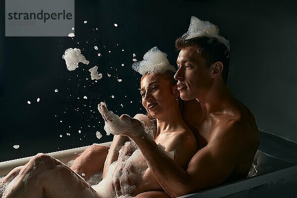 Seitenansicht eines jungen Mannes ohne Hemd und einer Frau mit geschlossenen Augen  die sich lächelnd berühren  während sie in einer Badewanne mit Schaum ruhen