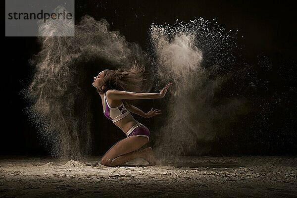 Junge sportliche Frau  die mit geschlossenen Augen den Sand vom Boden aufwirft  vor schwarzem Hintergrund