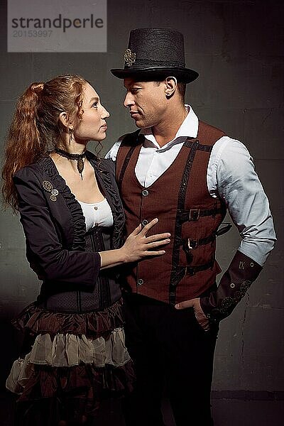 Junge rothaarige Frau und Mann in Vintage Steampunk Kostümen stehen und schauen sich gegenseitig vor grauem Hintergrund an