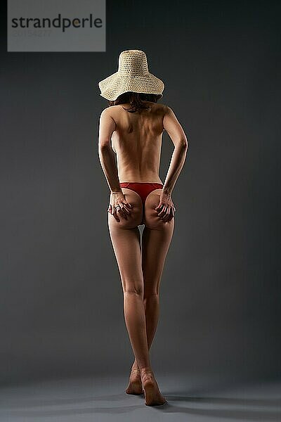 Ganzkörper Rückenansicht von sexy schlanke nackte Frau trägt rote Höschen und Strohhut stehen auf Zehenspitzen im Studio gegen grauen Hintergrund