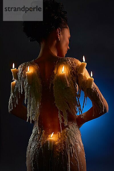 Rückenansicht eines jungen nackten weiblichen Modells mit brennenden Kerzen und Wachs  das den nackten Körper in blauer Dunkelheit bedeckt