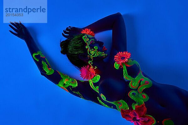 Draufsicht auf nacktes weibliches Modell mit nackter Brust und grüner fluoreszierender Farbe auf der Haut vor blauem Hintergrund liegend Neon Body Art Konzept