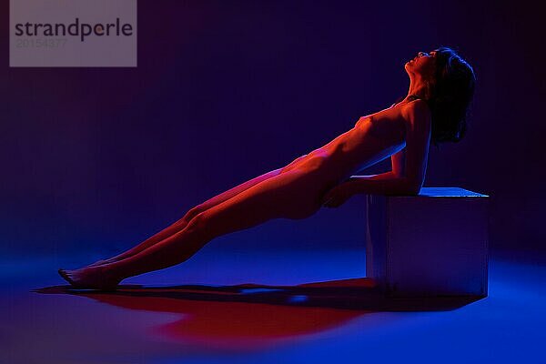 Ganzkörper Seitenansicht eines jungen nackten weiblichen Modells  das sich in einem dunklen  von blauen und roten Neonröhren beleuchteten Studio auf einen Würfel stützt
