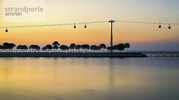 Silhouetten von Bäumen und Seilbahnen vor einem heiteren Sonnenuntergang  der sich auf dem ruhigen Wasser spiegelt  Lissabon