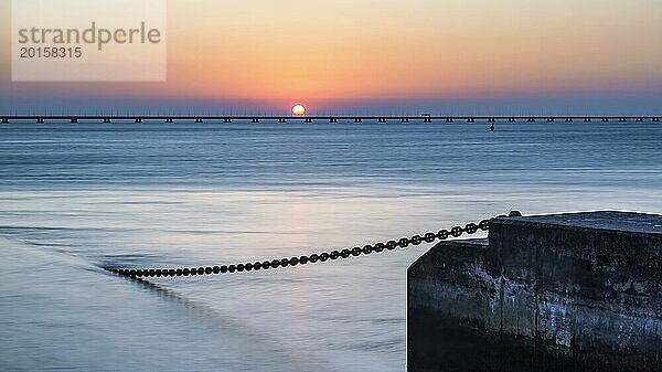 Ein heiterer Sonnenuntergang mit der Sonne  die den Horizont berührt  neben einem ruhigen Meer und einem Pier  der sich in die Ferne erstreckt  Lissabon