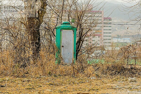 Grüne tragbare Toilette mit schmutziger weißer Tür vor kahlen  blattlosen Bäumen mit Gebäuden im Hintergrund in Südkorea