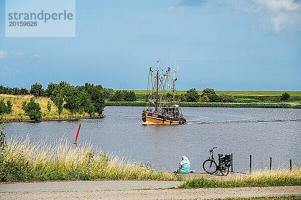 Ein Fischerboot fährt auf einem ruhigen Fluss vorbei  während eine Person neben einem Fahrrad sitzt und zuschaut  Fischkutter  Greetsiel  Norden  Ostfriesland  Niedersachsen