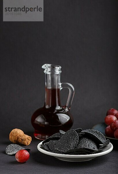 Schwarze Kartoffelchips mit Holzkohle  Balsamico Essig im Glas  rote Trauben auf einem blauen Keramikteller auf schwarzem Hintergrund. Seitenansicht  Nahaufnahme  selektiver Fokus  Kopierraum