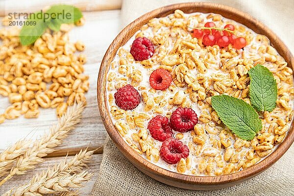 Weizenflockenbrei mit Milch  Himbeeren und Johannisbeeren in einer Holzschale auf weißem Holzhintergrund und Leinentextil. Seitenansicht  Nahaufnahme  selektiver Fokus