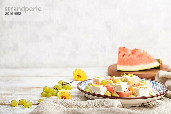 Vegetarischer Salat mit Wassermelone  Fetakäse und Trauben auf blauem Keramikteller auf weißem Holzhintergrund und Leinentuch. Seitenansicht  Kopierraum  selektiver Fokus