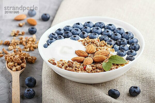 Joghurt mit Blaubeeren  Müsli und Mandeln in weißem Teller mit Holzlöffel auf grauem Betonhintergrund und Leinenstoff. Seitenansicht  Nahaufnahme  selektiver Fokus