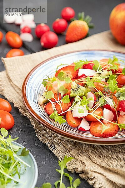 Vegetarischer Obst und Gemüsesalat aus Erdbeeren  Kiwi  Tomaten  Mikrogrünsprossen auf schwarzem Betonhintergrund und Leinentextil. Seitenansicht  Nahaufnahme  selektiver Fokus