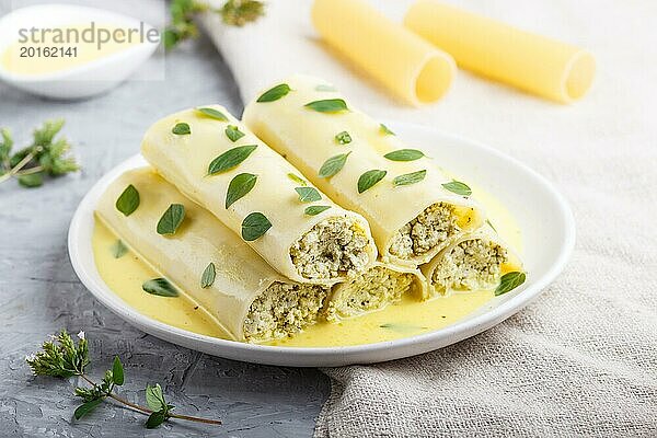 Cannelloni Nudeln mit Eiersoße  Frischkäse und Oreganoblättern auf grauem Betonhintergrund mit Leinenstoff. Seitenansicht  Nahaufnahme  selektiver Fokus