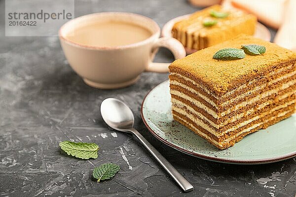 Hausgemachter Honigkuchen mit Milchcreme und Minze mit einer Tasse Kaffee auf einem schwarzen Betonhintergrund und orangefarbenem Textil. Seitenansicht  Nahaufnahme  selektiver Fokus