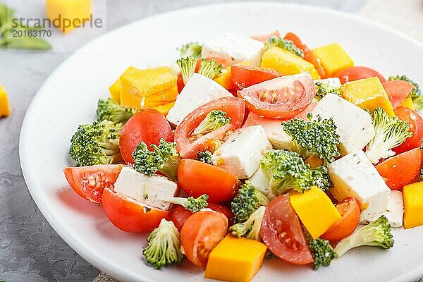 Vegetarischer Salat mit Brokkoli  Tomaten  Fetakäse und Kürbis auf weißem Keramikteller auf grauem Betonhintergrund und Leinentuch  Seitenansicht  Nahaufnahme  selektiver Fokus