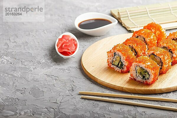 Japanische Maki Sushi Rollen mit fliegendem Fischrogen  Essstäbchen  Sojasauce und mariniertem Ingwer auf einem Holzbrett auf grauem Betonhintergrund. Seitenansicht  Kopierraum  selektiver Fokus