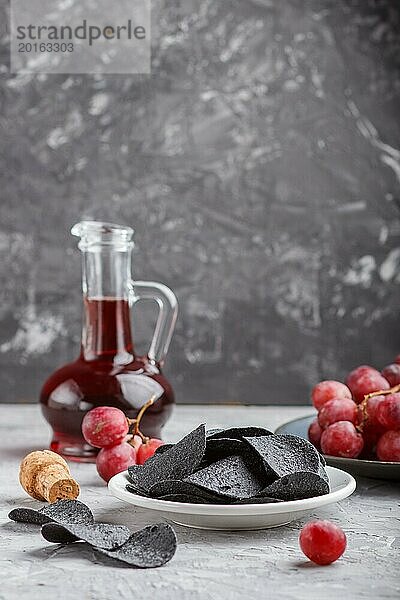 Schwarze Kartoffelchips mit Holzkohle  Balsamico Essig im Glas  rote Trauben auf einem blauen Keramikteller auf einem grauen Betonhintergrund. Seitenansicht  Nahaufnahme  selektiver Fokus  Kopierraum