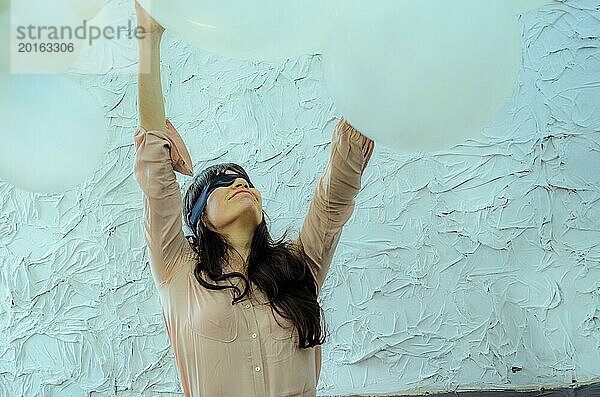 Eine spielerische junge erwachsene hispanische Frau mit verbundenen Augen streckt sich in einer heiteren Geste zwischen zwei großen Luftballons in die Höhe  künstlerische Performance  spielerischer Tanz